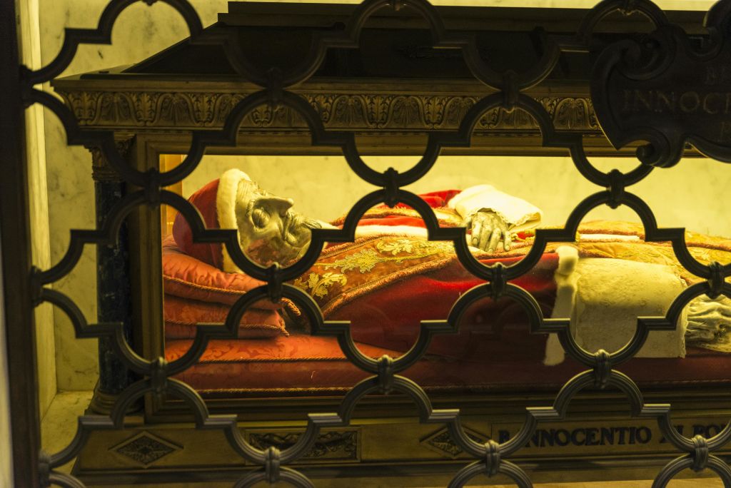 화려한 빨간색과 금색 가운을 입은 보존된 인물은 정교하게 디자인된 금속 그릴을 통해 볼 수 있는 유리 케이스 안에 있습니다.