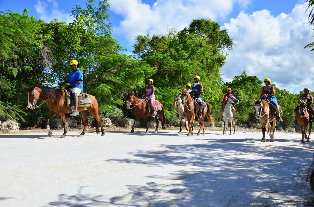 부분적으로 흐린 하늘 아래 녹지로 둘러싸인 흙길에서 말을 타고 헬멧을 쓴 사람들.