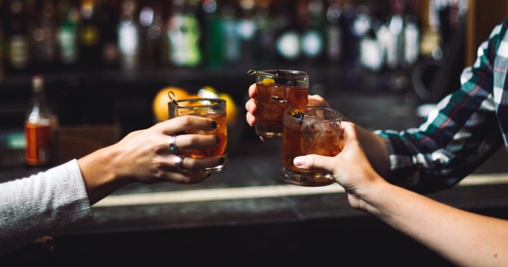 세 손이 토스트 제스처로 칵테일 잔을 들고 있습니다. 흐릿한 바 배경에 병이 보입니다.