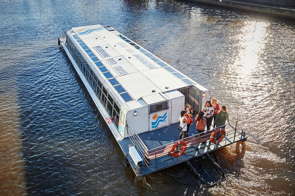 밝은 흰색 지붕과 안전 장비가 보이는 후면 데크에 사람들이 모인 강가의 관광 보트.