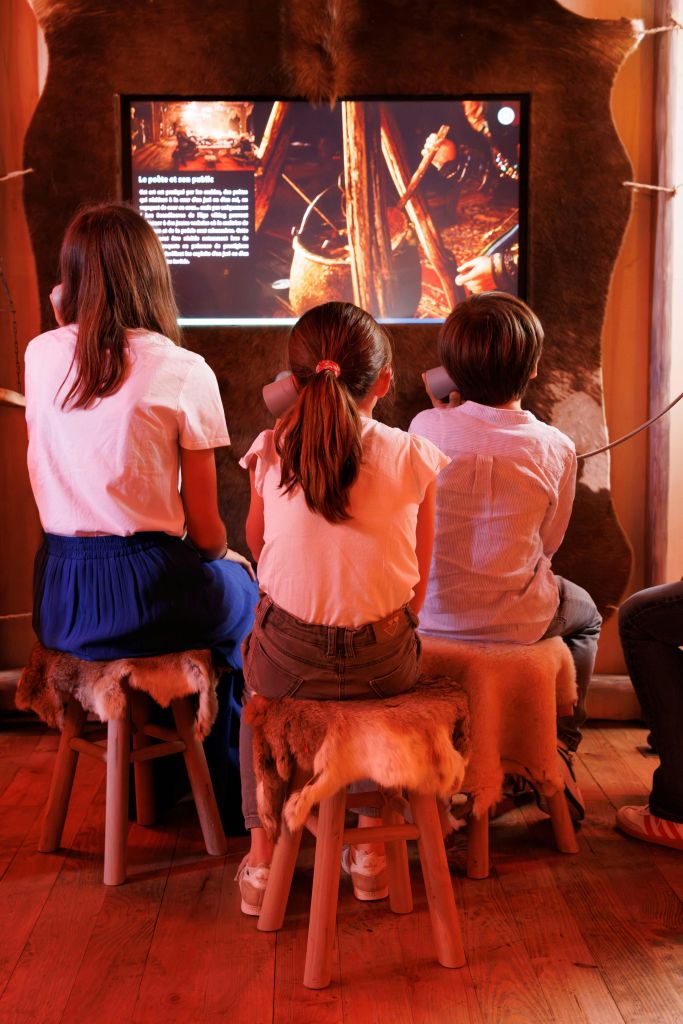 세 명의 어린이가 동물 가죽 커버가 달린 의자에 앉아 조명이 켜진 전시물을 보고 어두운 방에서 헤드폰으로 듣고 있습니다.