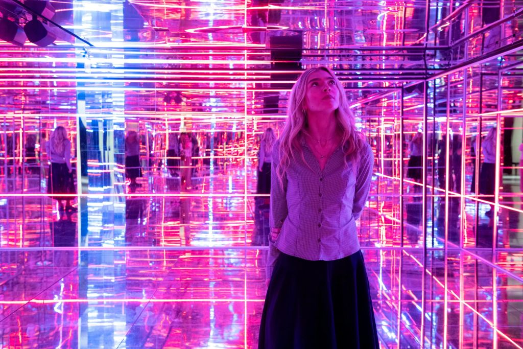 거울이 가득한 방에 선명한 분홍색과 보라색 네온 불빛이 비추는 한 여성이 서서 위를 바라보고 있습니다.