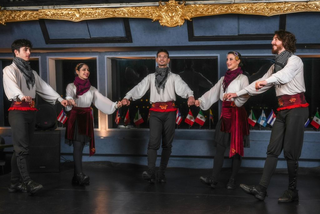 전통 무용복을 입은 네 사람이 손을 잡고 미소를 짓고 있으며, 배경에는 여러 개의 깃발이 있습니다.