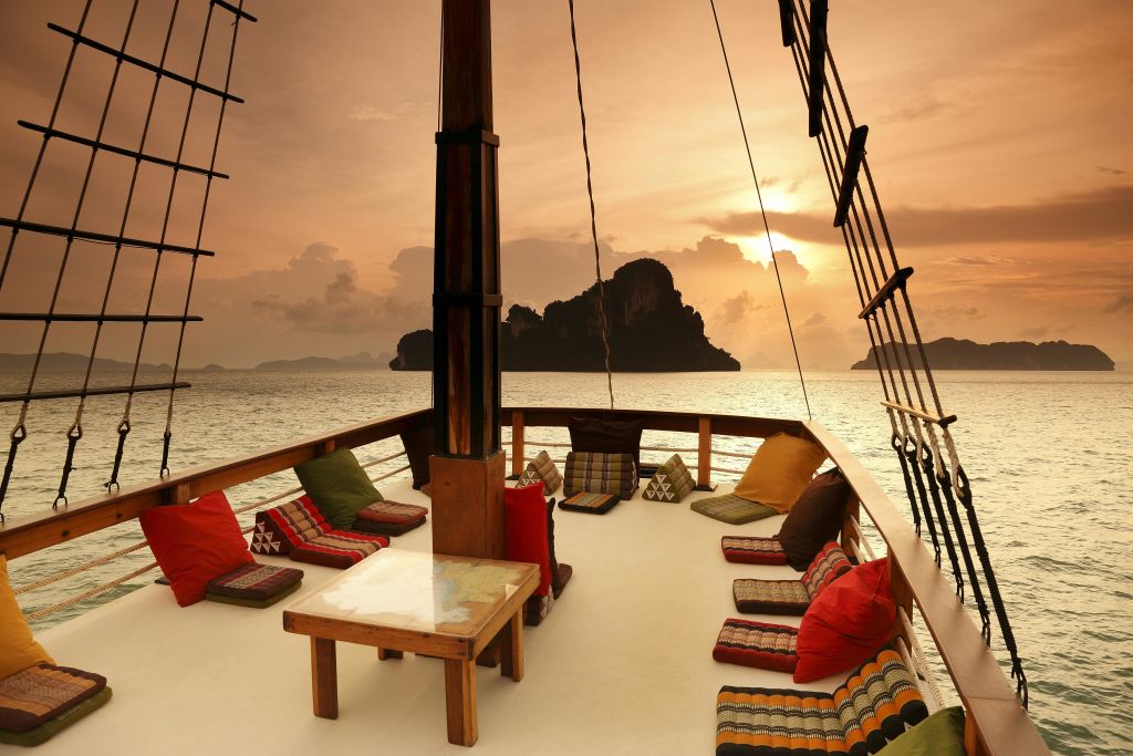 탁자 주위에 화려한 쿠션이 있는 보트 갑판으로, 해가 질 무렵 바위섬을 향해 항해합니다.