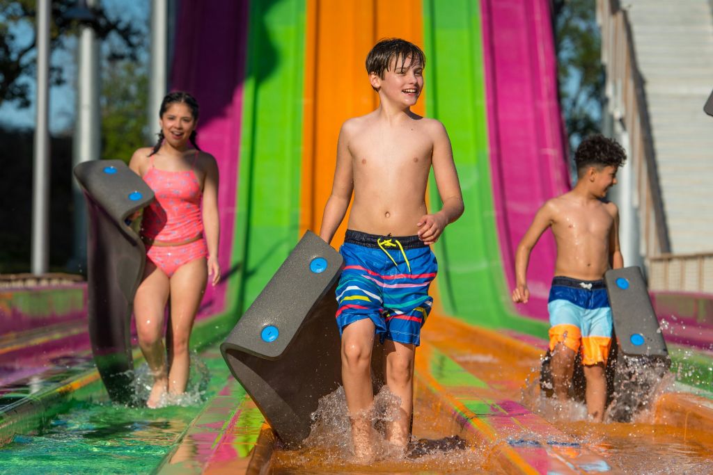 수영복을 입은 두 명의 어린이가 화창한 날 형형색색의 워터 슬라이드 레인을 걷는 동안 매트를 들고 다닙니다.