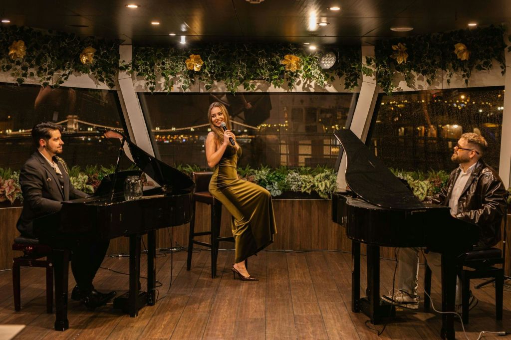 초록색 드레스를 입은 여성이 우아한 인테리어 디자인 아래 그랜드 피아노 두 대가 있는 무대에서 마이크를 잡으며 노래하고 있습니다.