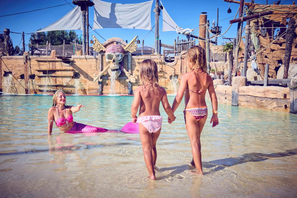 해적을 주제로 한 배경 근처에서 두 어린이가 인어공주 복장을 한 연기자와 마주보며 얕은 물가에 서 있습니다.