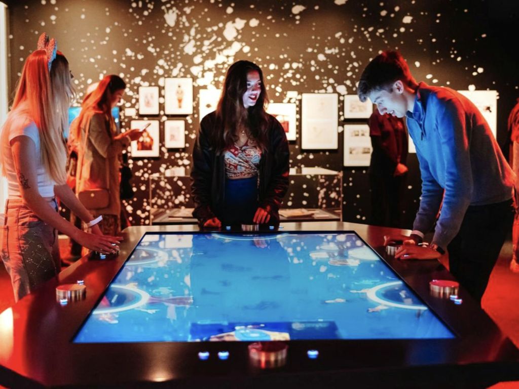 예술로 장식되고 푸른 빛이 비추는 어두운 방에서 커다란 대화형 터치스크린 테이블과 상호 작용하는 사람들.