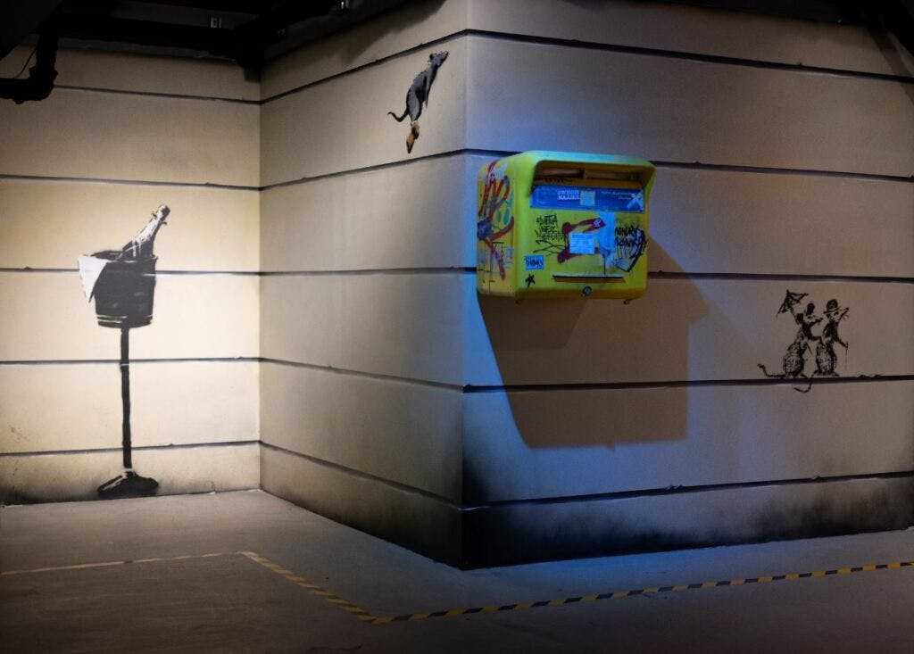 병과 쥐에서 액체를 붓는 남성이 있는 벽면 예술이 있는 방 구석. 벽에 붙어 있는 노란색 우편함.