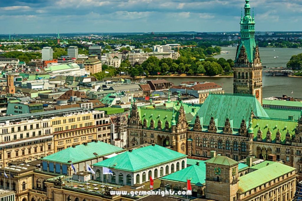 유서 깊은 건물, 초록색 옥상, 높은 탑, 부분적으로 흐린 하늘 아래 강을 배경으로 도시 풍경을 조망할 수 있습니다.