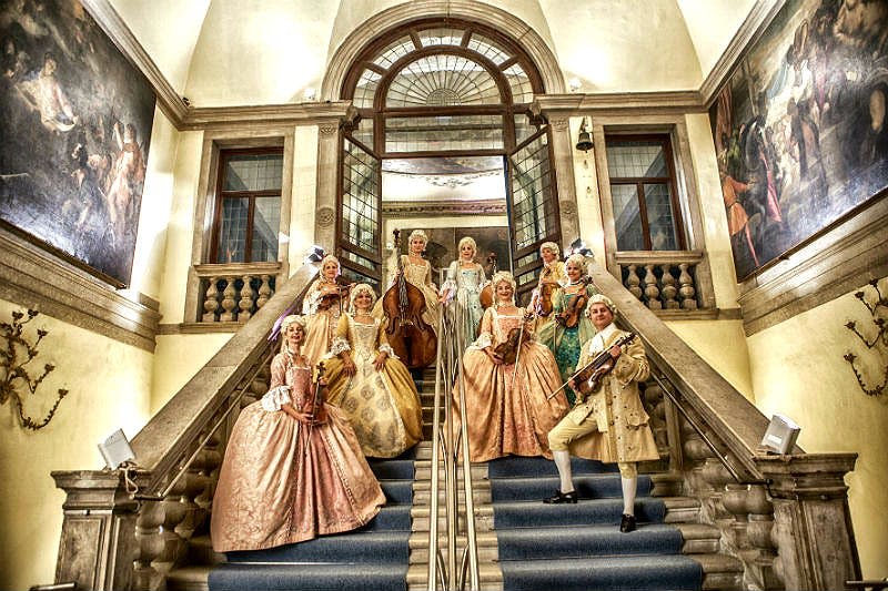 정교한 의상을 입고 악기를 들고 호화로운 건물의 웅장한 계단에 서 있는 사람들의 그룹.