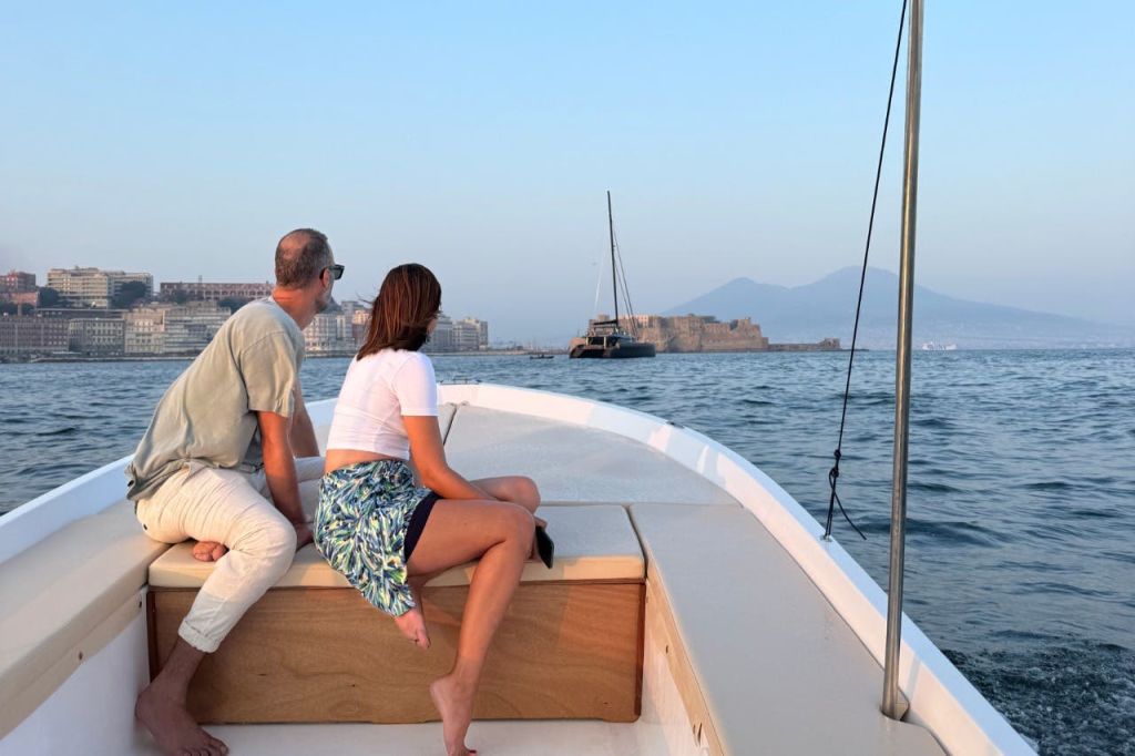 두 사람이 보트 갑판에 앉아 잔잔한 바다 너머로 멀리 떨어진 도시 풍경과 산을 바라보고 있습니다.