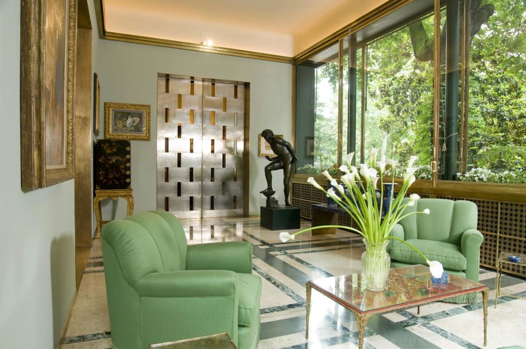 초록색 안락의자가 있는 객실, 흰색 꽃이 있는 유리 커피 테이블, 커다란 창문, 동상, 금속으로 포인트를 준 벽이 있습니다.