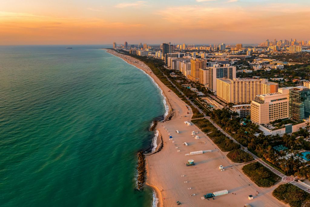 부분적으로 흐린 하늘 아래 베이지색 모래 해변, 청록색 물, 수많은 고층 건물이 있는 해안선의 공중 전망.