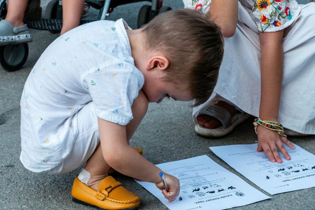 성인의 도움을 받아 인쇄된 워크시트에 무릎을 꿇고 글을 쓰는 어린이. 둘 다 야외에 있으며, 아이에게 초점을 맞춥니다.
