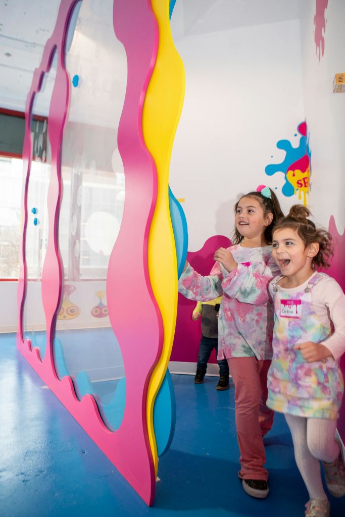 화려한 색상의 셔츠를 입은 두 어린이가 화사하게 꾸며진 방의 분홍색과 노란색 전시관 근처에서 미소를 짓고 놀고 있습니다.