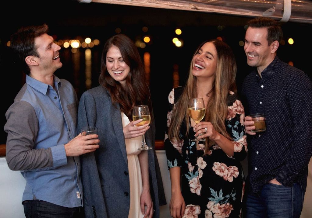 미소를 짓는 세 사람이 음료를 들고 하룻밤을 즐깁니다. 배경이 불빛으로 흐릿해졌다. 캐주얼한 차림의 남성 한 명, 여성 두 명.