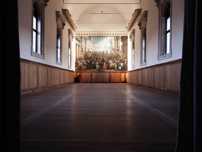 나무 바닥과 벽 패널이 있는 크고 빈 방으로, 먼 벽에 디테일한 벽화가 있습니다.