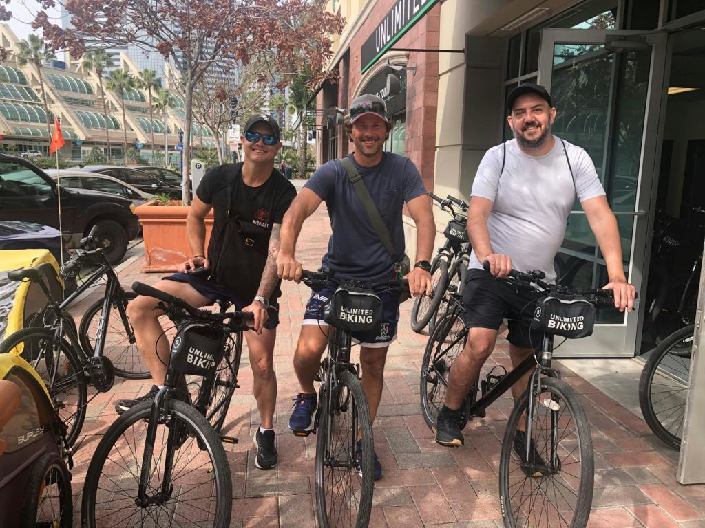 '언리미티드 바이킹 (Unlimited Biking)' 이라는 이름의 자전거 대여점 밖에서 세 사람이 자전거를 타고 화창한 날 라이딩을 준비하며 미소를 짓고 있습니다.