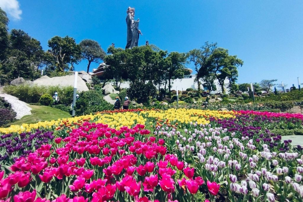 다양한 색상의 활기찬 튤립이 있는 정원과 맑고 푸른 하늘 아래 울창한 나무로 둘러싸인 높은 동상을 배경으로 합니다.