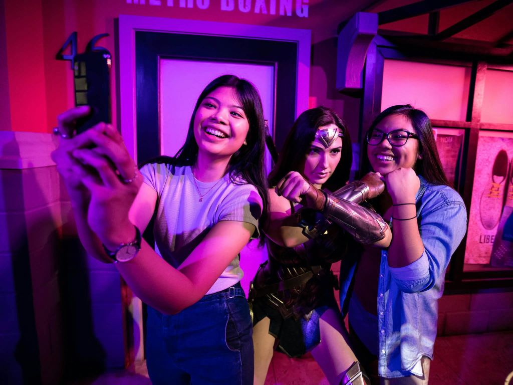 활기찬 조명이 있는 방 안의 원더우먼 동상 옆에서 미소 짓는 두 여성이 포즈를 취하고 있습니다.