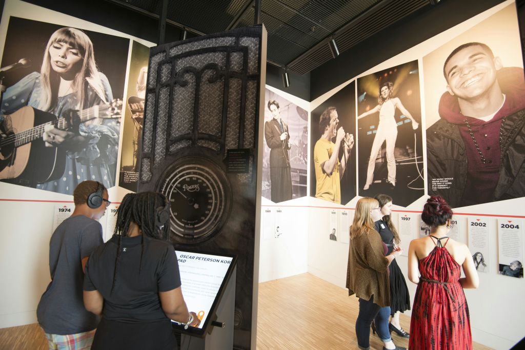 헤드폰을 착용한 두 사람이 박물관 전시물과 상호작용합니다. 음악가의 대형 사진이 배경의 벽에 표시됩니다.