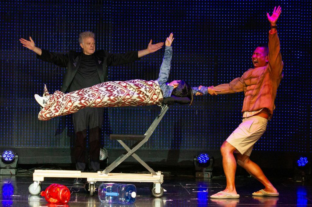 공연자가 무대에서 무늬가 있는 담요로 감싼 사람을 공중 부양하고 있으며, 주변에 조명과 소품이 보입니다.