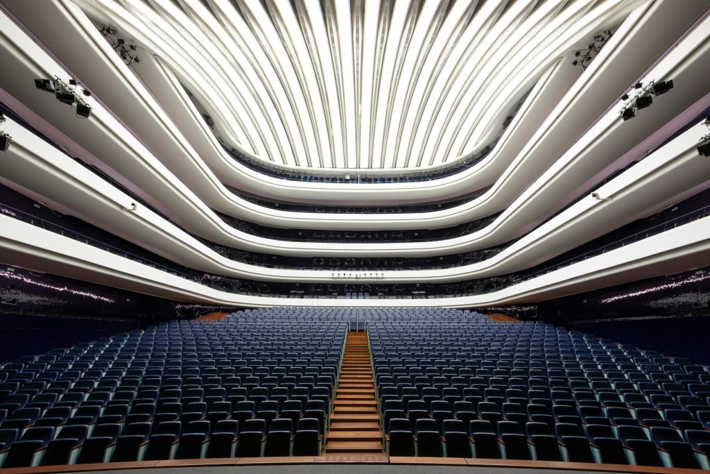 여러 개의 곡선형 발코니, 중앙 계단, 밝고 늑골이 있는 천장 아래 파란색 좌석이 줄지어 있는 텅 빈 현대식 강당입니다.