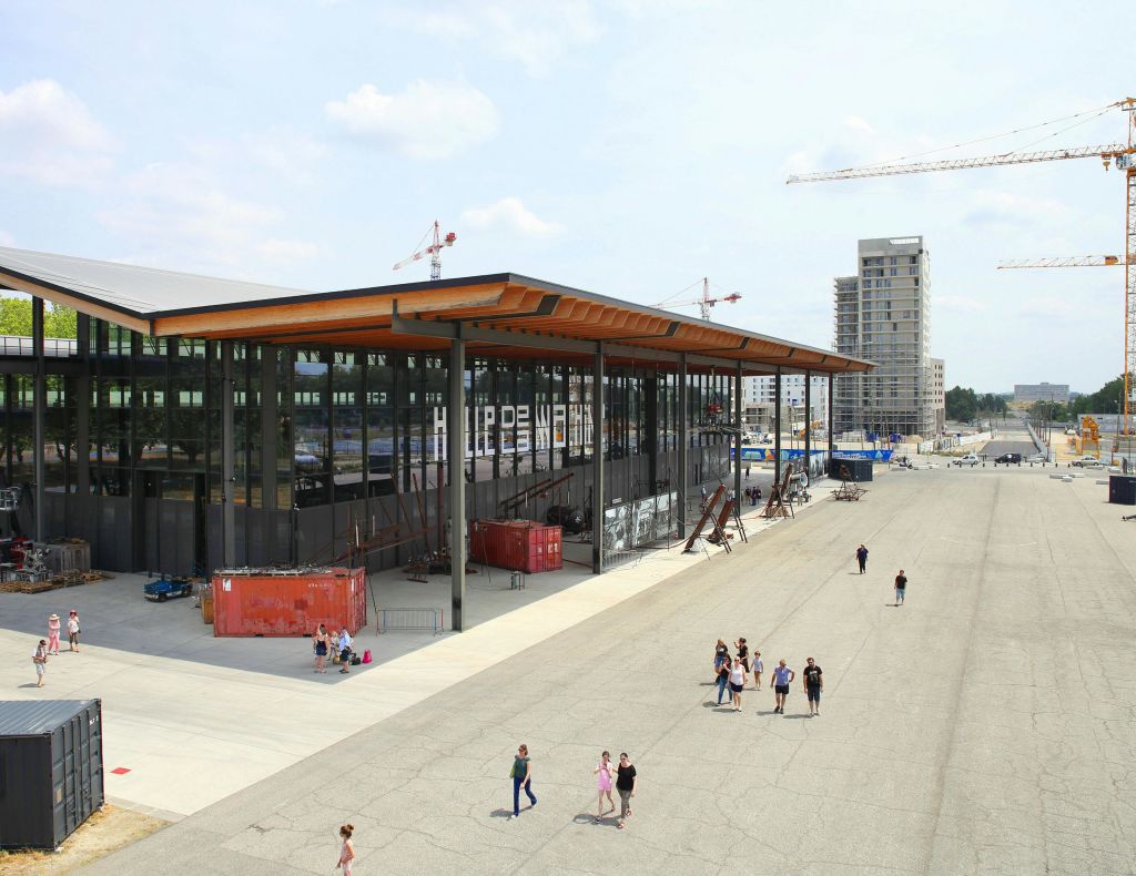 사람들은 크레인과 고층 건물을 배경으로 현대적이고 유리로 덮인 건물 옆의 넓은 보도를 걷습니다.