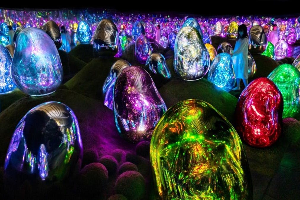 어둡고 잔디가 우거진 풍경에 커다랗고 빛나는 여러 가지 빛깔의 달걀 모양의 조형물 사이를 걷는 사람들.