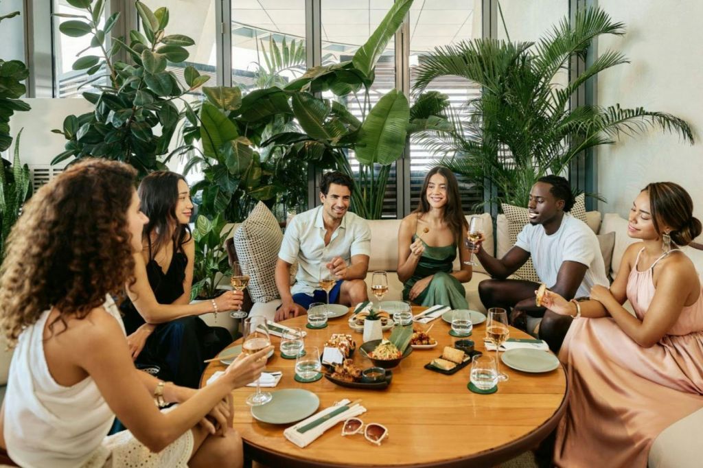 5명이 우거진 실내 식물로 둘러싸인 나무 테이블에 앉아 음식과 음료를 즐기고 있습니다. 그들은 웃으며 이야기하고 있습니다.