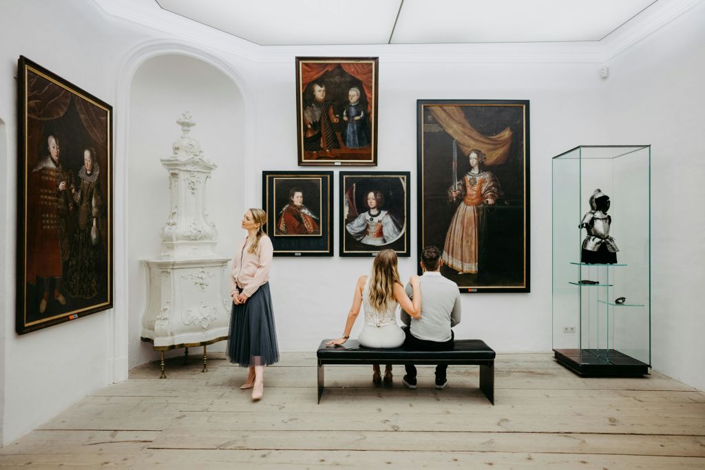 한 커플이 미술관의 벤치에 앉아 초상화를 감상하고 있고, 한 여성이 근처에 서서 주변을 관찰하고 있습니다.