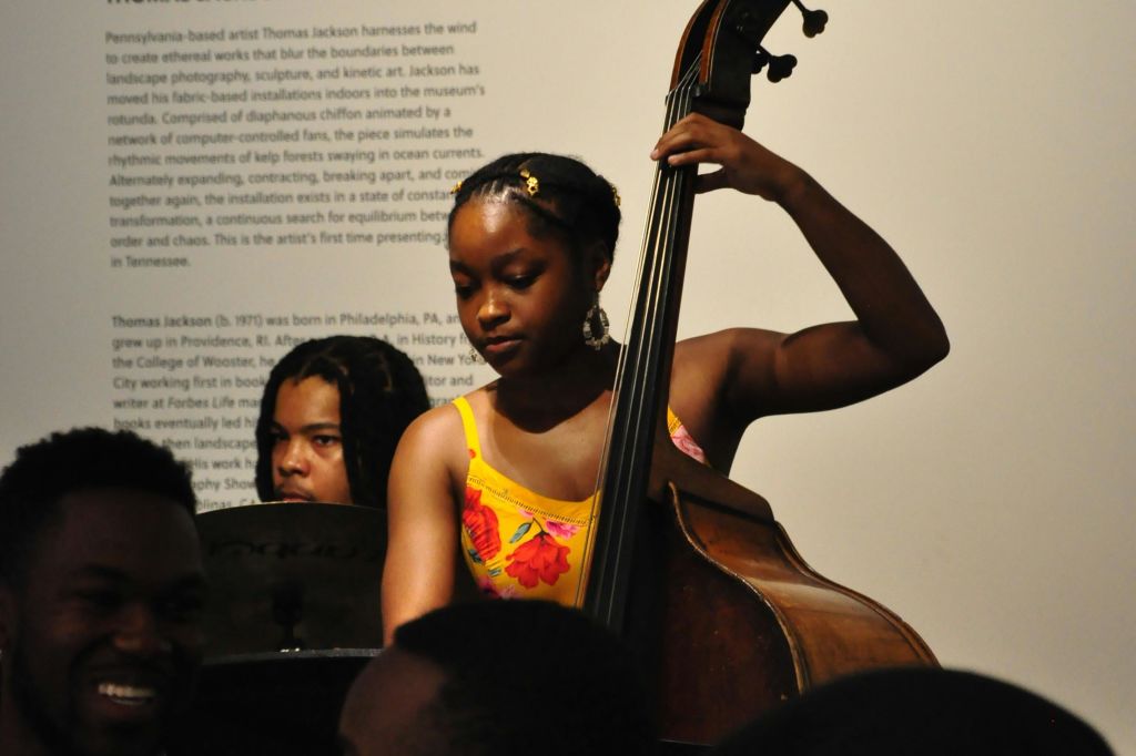 노란색 꽃무늬 드레스를 입은 젊은 여성이 더블 베이스를 연주하고 드러머가 배경으로 등장합니다. 텍스트가 있는 벽이 부분적으로 보입니다.