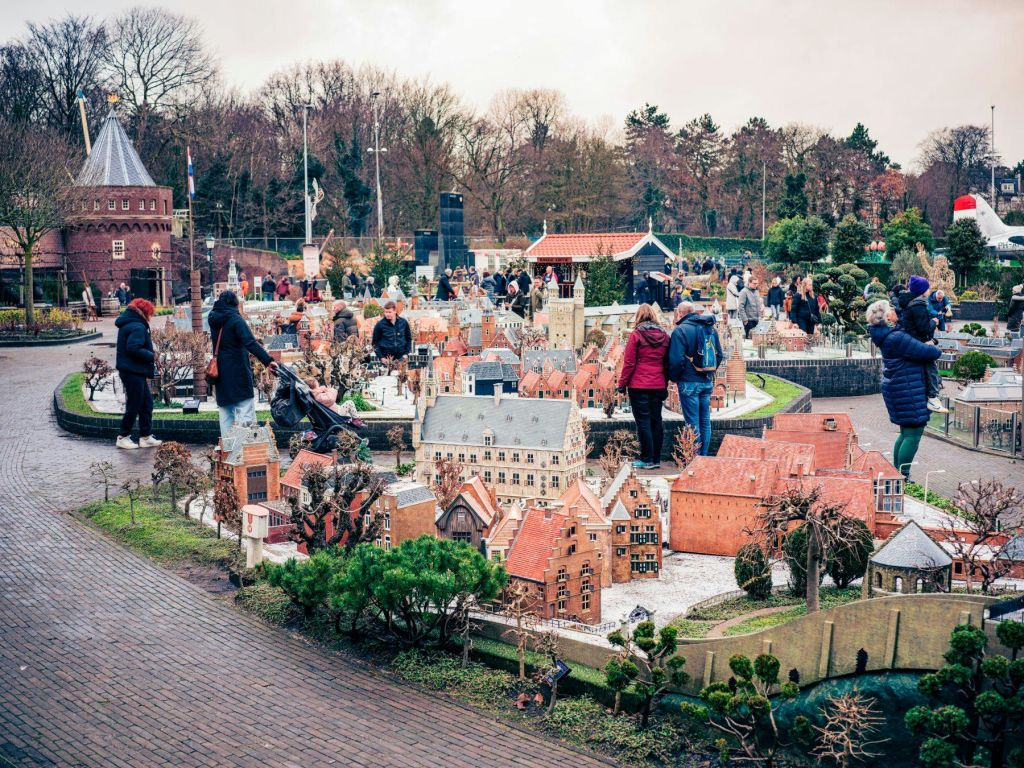 숲이 우거진 배경을 배경으로 디테일한 건물과 나무가 있는 야외 미니어처 도시 전광판을 둘러보는 사람들이 많습니다.