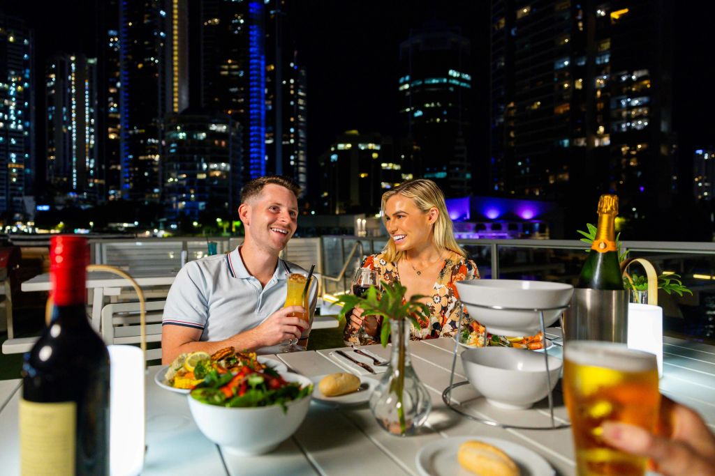 도시의 불빛과 고층 건물로 둘러싸인 야외 레스토랑에서 저녁 식사를 즐기는 커플. 테이블 위에 음료와 음식이 놓여 있습니다.