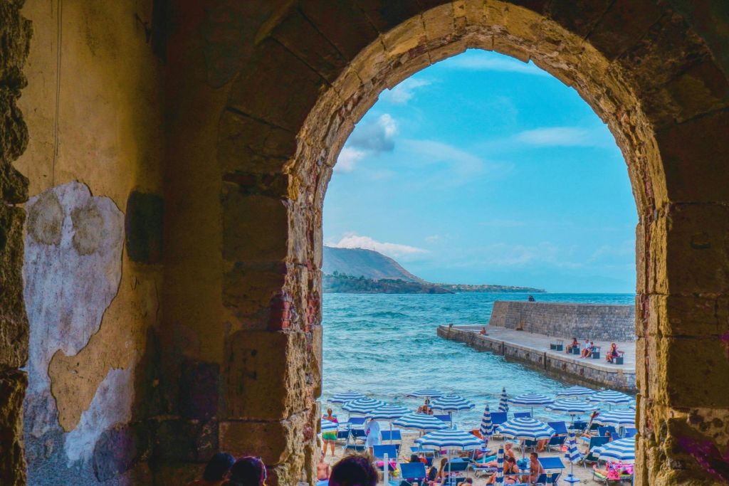 푸른 우산을 쓴 해변의 석조 아치, 일광욕을 즐기는 사람들, 바다로 뻗은 선착장, 저 멀리 산이 보이는 풍경.