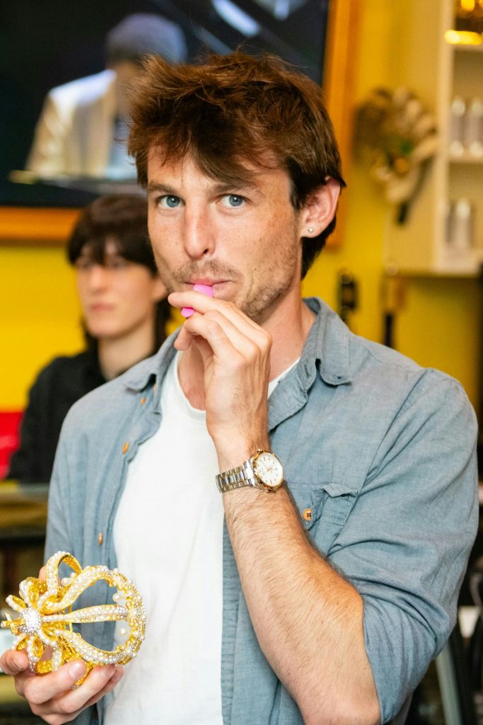 짧은 머리카락을 가진 사람이 노란색 벽과 장식이 있는 캐주얼한 실내 환경에서 입 근처에 분홍색 물건을 들고 있습니다.