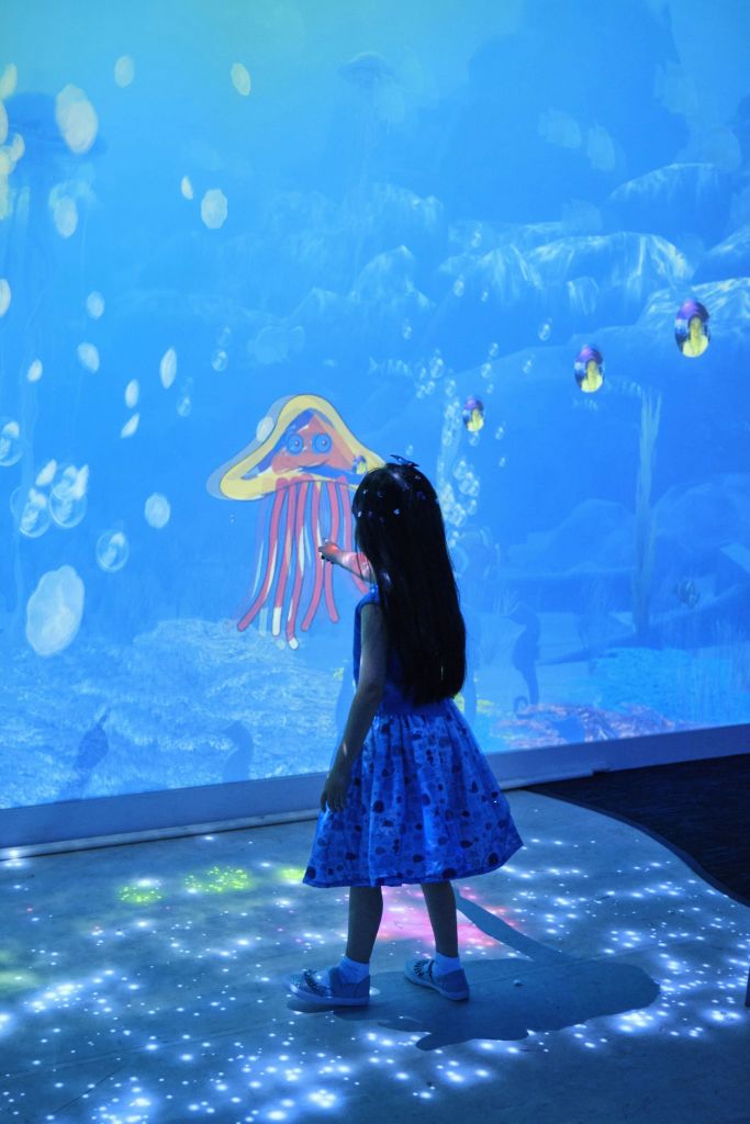 파란색 드레스를 입은 어린이가 해파리와 거품이 있는 수중 장면을 보여주는 빛나는 화면을 터치합니다.