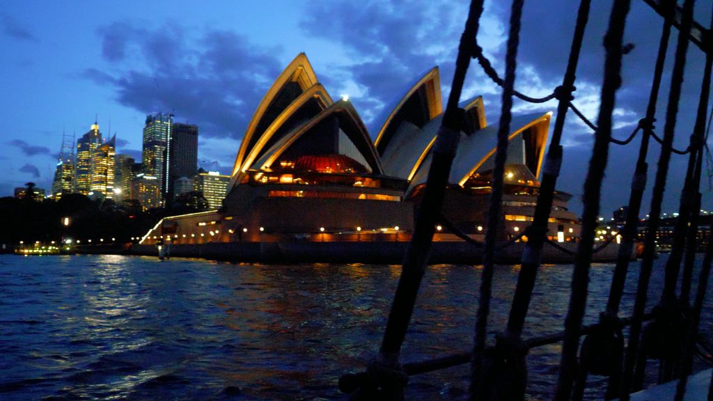 해질 무렵 밤하늘을 배경으로 조명이 켜진 시드니 오페라하우스의 전경. 전경에 물과 보트의 일부가 있습니다.