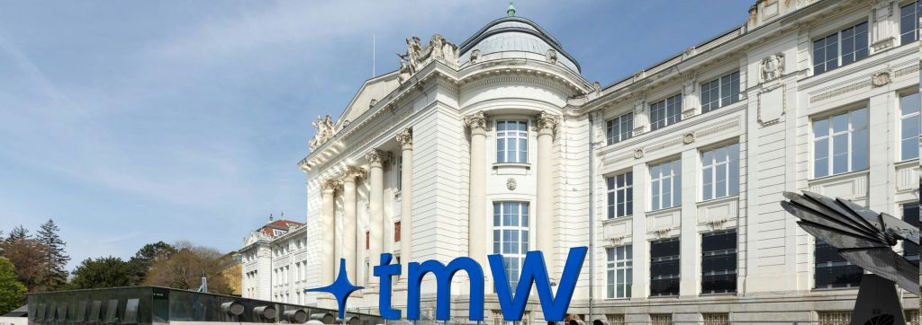 입구 위에 높은 기둥과 조각이 있는 화려한 흰색 건물, 전면에 커다란 파란색 'tmW' 표지판이 있습니다.