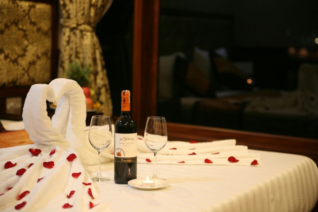 와인 한 병, 와인 잔 두 개, 장미 꽃잎, 테이블 위에 불이 켜진 촛불이 놓인 로맨틱한 공간입니다. 백그라운드에 도톰한 수건이 있습니다.