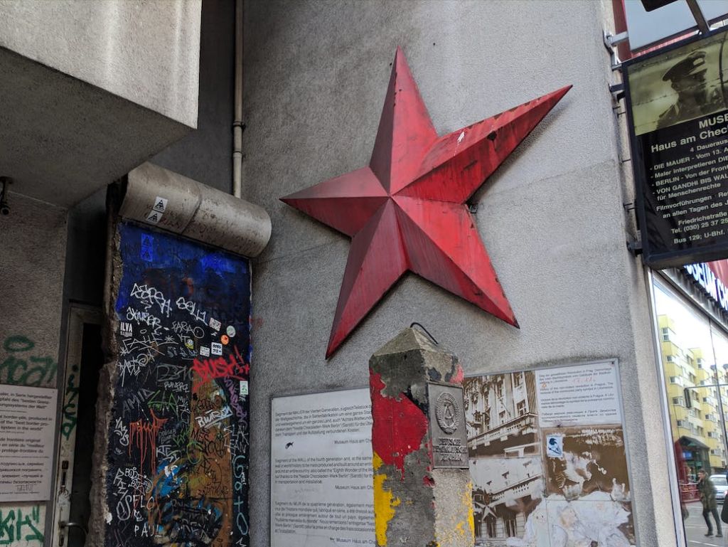 회색 벽에 있는 커다란 붉은 별, 인접한 표면에 있는 화려한 낙서, 정보용 플라크가 있는 풍화 기념물.