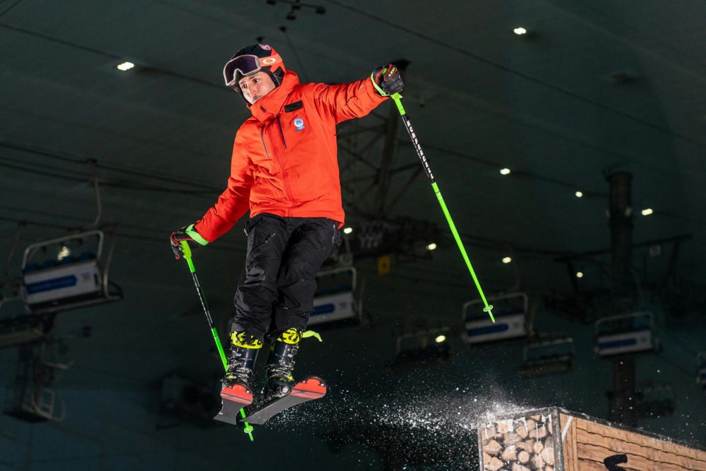 오렌지색 재킷과 스키 장비를 입은 사람이 실내에서 스키를 타면서 녹색 스키 폴을 들고 있습니다.