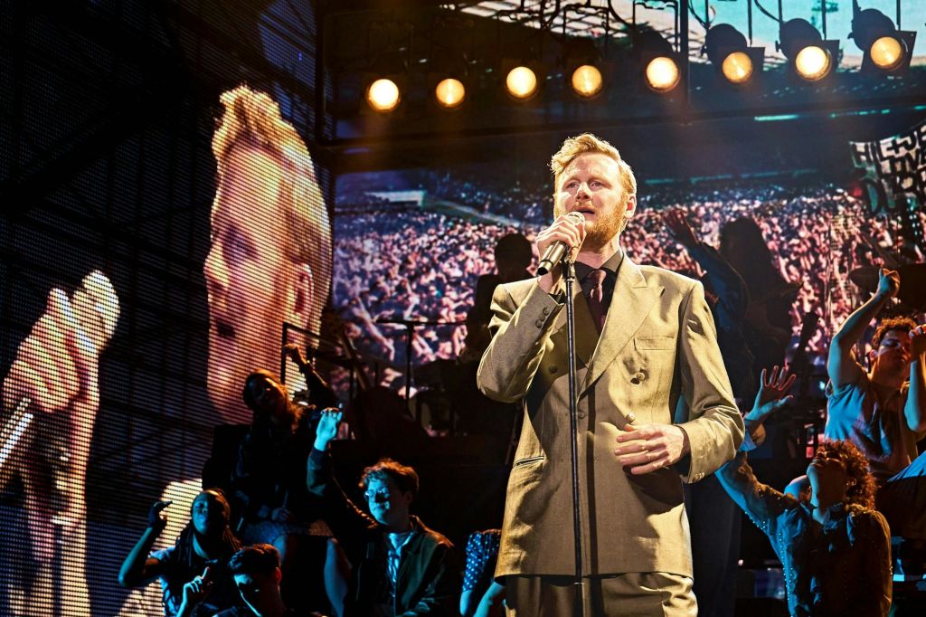 무대에서 양복을 입은 한 남성이 무대 뒤 스크린에 큰 무리가 있고 주변 사람들이 마이크를 잡고 노래하고 있습니다.