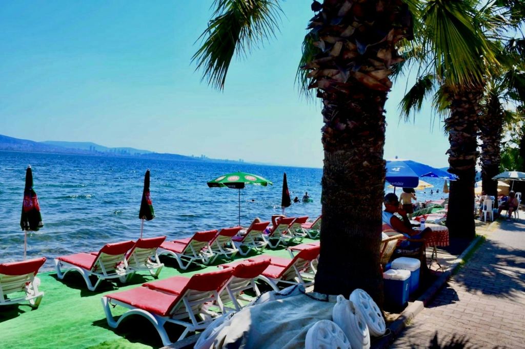 파라솔 아래 바다가 보이는 라운지 의자가 있는 해변 풍경. 손바닥과 사람이 배경으로 보입니다.