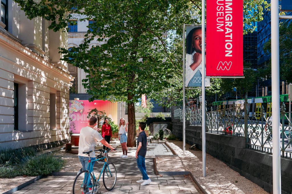 빨간색과 흰색 박물관 배너 옆에 있는 이민 박물관 외부의 가로수 길 근처에서 걷고 자전거를 타는 사람들.