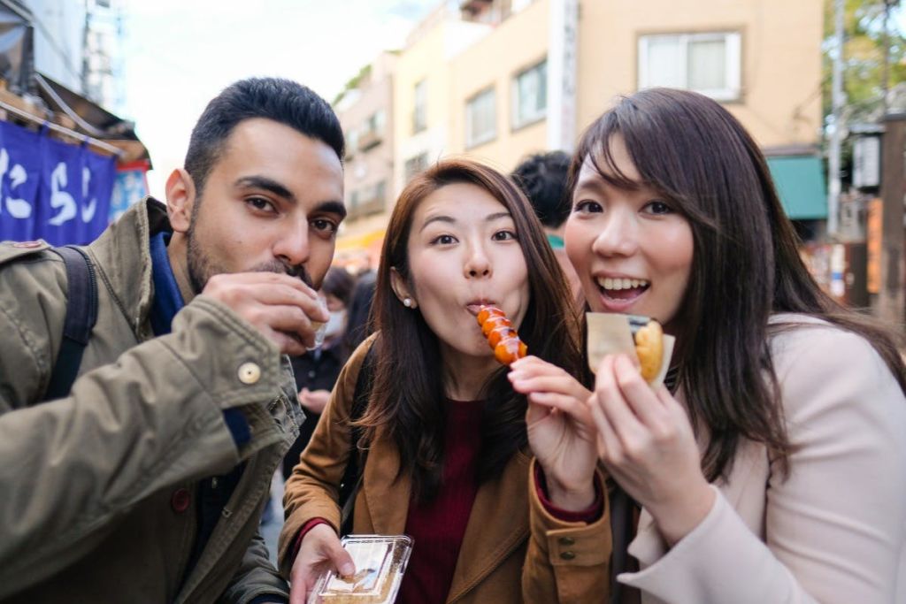 두 여성이 길거리 음식을 즐기며 웃고 있으며, 한 여성은 꼬치를 들고 다른 한 여성은 샌드위치를 들고 있습니다. 도시를 배경으로 한 공간.
