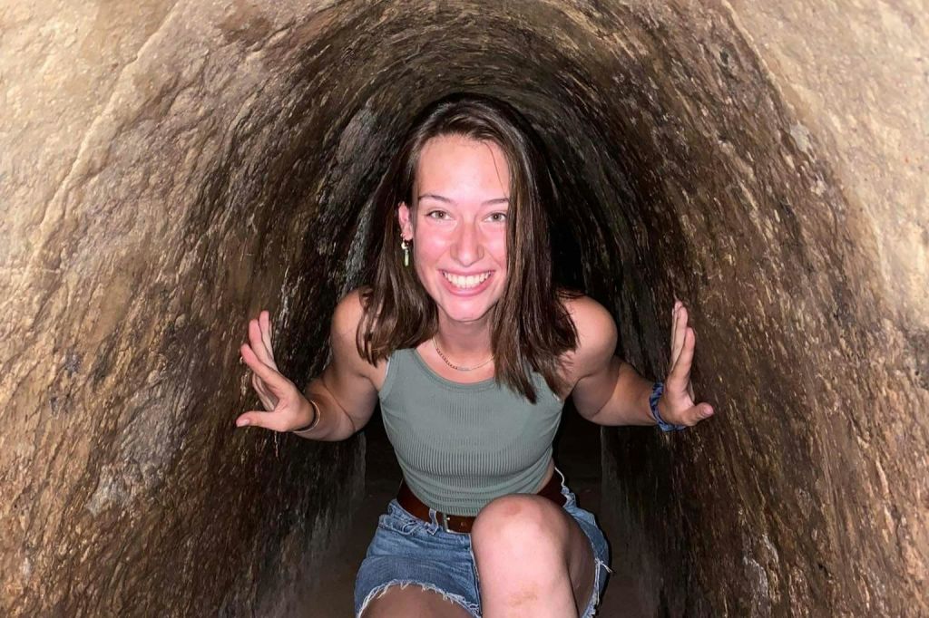 한 사람이 좁고 바위 같은 터널 안에 웅크리고 앉아 카메라를 보며 미소를 짓고 녹색 탱크탑과 데님 반바지를 입고 있습니다.