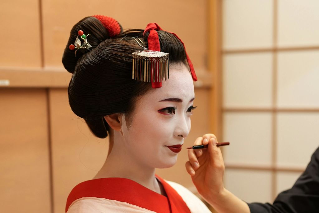 정교한 헤어 장식과 하얀 얼굴 메이크업을 한 전통 의상을 입은 여성이 조심스럽게 입술을 칠하고 있습니다.