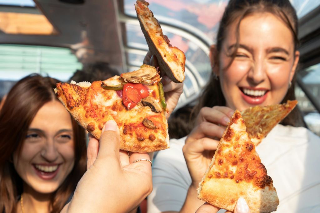 피자 조각을 들고 웃고 있는 사람들. 조각 중 하나에 버섯과 고추 같은 토핑이 보입니다.
